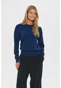 Frau mit langen blonden Haaren, die einen dunkelblauen Pullover und schwarze Hosen trägt, steht mit den Händen in den Taschen vor einem einfarbigen Hintergrund.