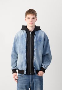 Chaqueta de mezclilla de lavado azul claro, con un frente abotonado y puños acanalados negros, superpuesta a una sudadera con cremallera negra.