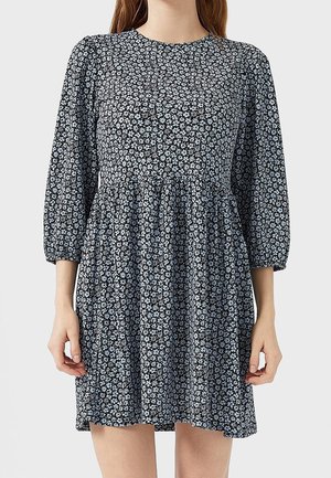 Femme portant une robe noire à petits motifs floraux blancs, manches trois-quarts bouffantes, et taille froncée, debout devant un fond uni.