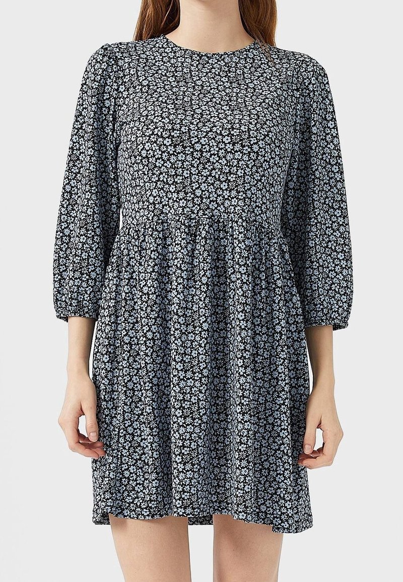 Femme portant une robe noire à petits motifs floraux blancs, manches trois-quarts bouffantes, et taille froncée, debout devant un fond uni.