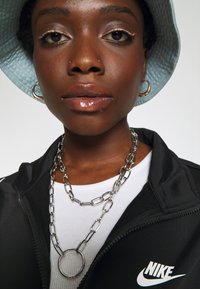 Giacca nera con zip e logo Nike, collana a catena d'argento sovrapposta, maglietta bianca a coste e cappello a secchiello blu chiaro. Labbra lucide e orecchini a cerchio.