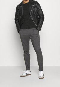 Czarna skórzana kurtka motocyklowa, ciemny dziany sweter, szare dopasowane spodnie oraz białe trampki z czarnymi paskami. Teksturowane materiały; nowoczesny krój.