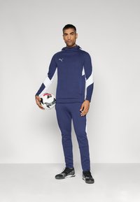Navyblauwe sportieve trainingsoutfit met witte zijpanelen, bestaande uit een hoodie en taps toelopende broek. Model houdt een voetbal vast.