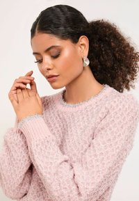 Világos rózsaszín pulóver textúrált, gyémántmintázattal és ezüstszálas díszítéssel a gallérnál és az ujjaknál. A modellen ezüst fülbevalók vannak.