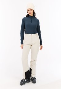 Licht beige skibroek met zwarte accenten, gecombineerd met een donkerblauwe lange mouw top en een crèmekleurige gebreide muts. Zwarte skischoe.