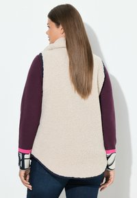 Faux-Shearling-Weste in Creme mit hohem Kragen, kombiniert mit einem burgunderfarbenen Langarmshirt und gemusterten Manschetten. Glatte und strukturierte Oberflächen.