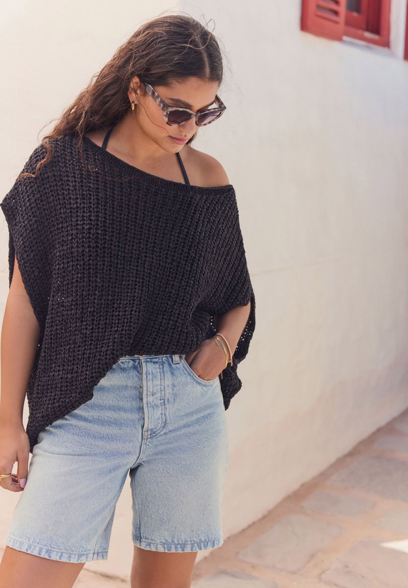 Maglione nero a maglia aperta con design scivolato sulle spalle, abbinato a pantaloncini di jeans azzurri chiari. Il look presenta una vestibilità rilassata e un contrasto di texture.