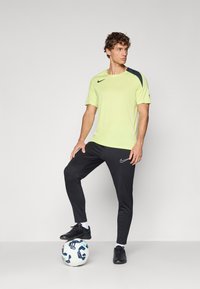 Camiseta deportiva Nike de color amarillo neón con acentos negros en los hombros, combinada con pantalones negros ajustados de Nike. El modelo está de pie sobre un balón de fútbol.