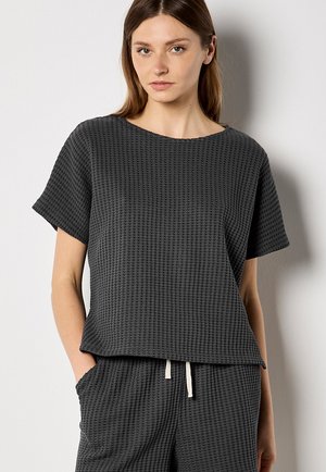 Kobieta w ciemnoszarym koszulce o teksturze wafla z krótkim rękawem i pasujących spodniach ze ściągaczem, stojąca z rękami w kieszeniach.