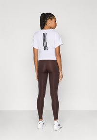Hvid t-shirt med korte ærmer og tre sorte striber på ryggen, kombineret med brune leggings og hvide sports sko.