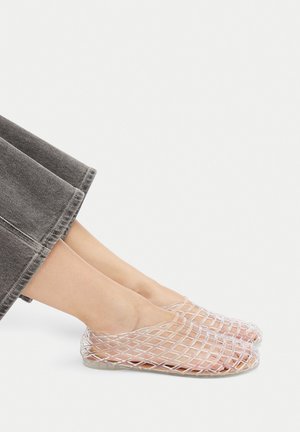 Gennemsigtige plastiksko med gitterdesign, åben tå og flad sål, sat mod gråt denimstof.
