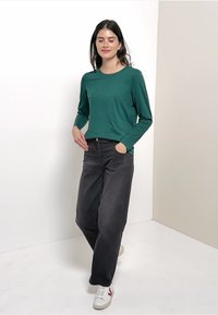 Grünes Langarmshirt kombiniert mit dunklen, locker sitzenden Jeans und weißen Turnschuhen. Einfaches Design ohne sichtbare Muster oder Verzierungen.