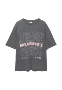 Šedé bavlněné tričko s oversize střihem. Na přední straně je opakovaný text "SAKAMOTO'S" tučným černým a růžovým písmem. Kulatý výstřih, krátké rukávy.