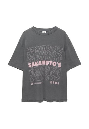 T-shirt en coton gris à coupe oversize. Le devant présente le texte répété "SAKAMOTO'S" en gras noir et rose. Encolure ronde, manches courtes.