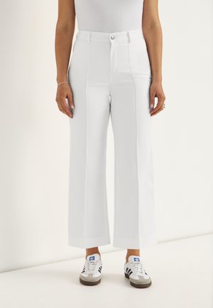 Witte, hoogwater, wijde broekspijpen gedragen met witte sneakers met blauwe strepen, te zien op een persoon die staat tegen een effen achtergrond.