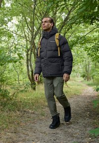 Svart puffy jakke med høy krage, khaki bukser og svarte tursko. Gul ryggsekk. Grønn skogssti med myk jord.