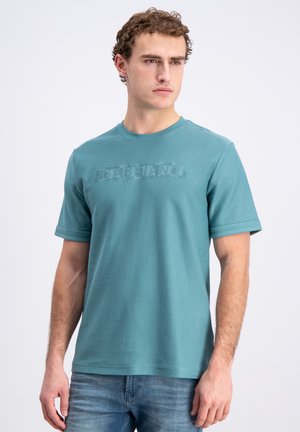 Jonge man met krullend haar die een teal korte mouw T-shirt met reliëftekst draagt, gecombineerd met een blauwe spijkerbroek, licht naar rechts kijkend.
