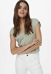 Top verde claro de manga corta con un sutil brillo, escote redondo y corte relajado, combinado con jeans blancos. Textura simple y suave.