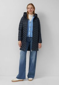 Cappotto imbottito blu navy con cappuccio, caratterizzato da cuciture a zigzag, abbinato a un cardigan azzurro chiaro e jeans in denim a zampa d'elefante. Sneakers bianche completano l'outfit.