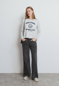 Mujer con camiseta de manga larga color gris claro con texto, vaqueros anchos negros, zapatillas blancas y un pequeño bolso negro de hombro.