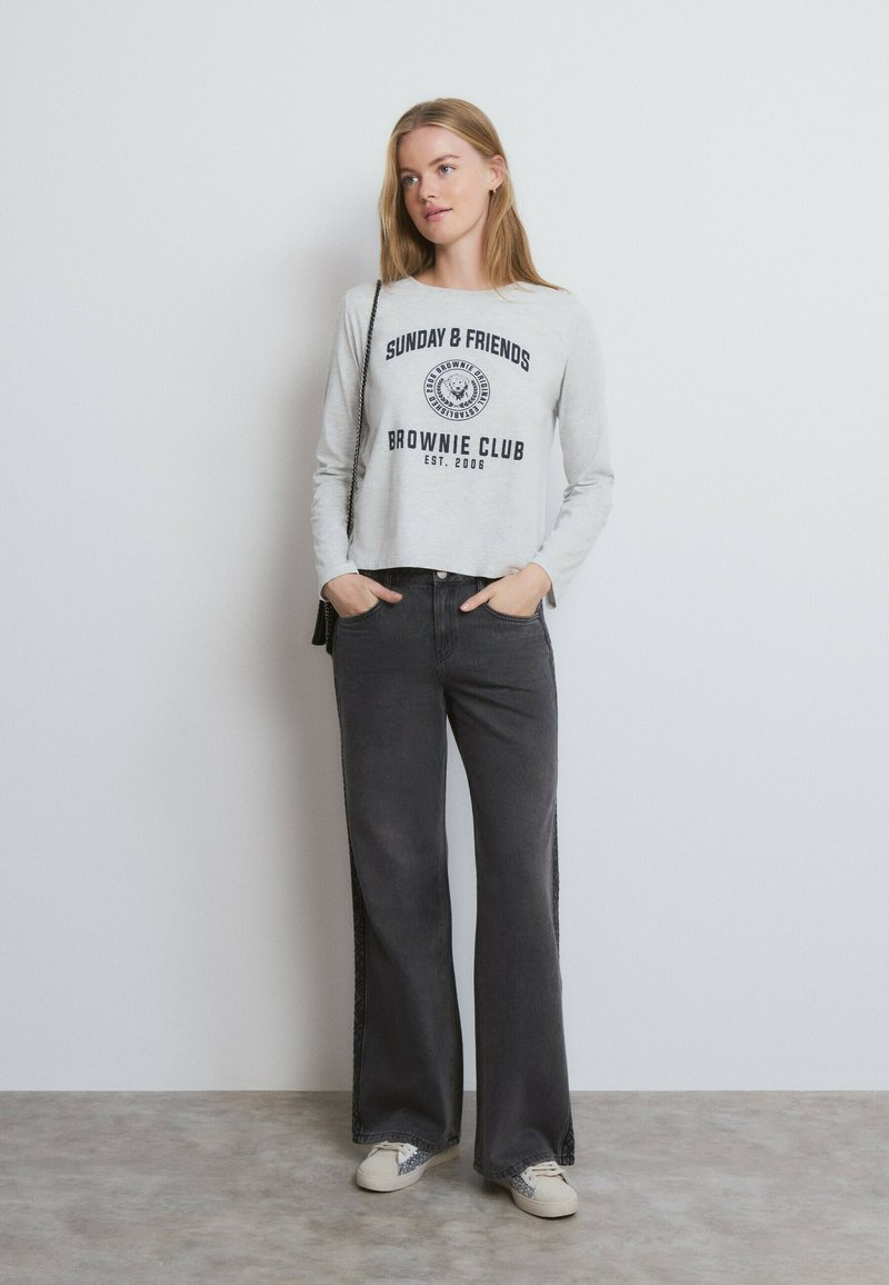 Mujer con camiseta de manga larga color gris claro con texto, vaqueros anchos negros, zapatillas blancas y un pequeño bolso negro de hombro.