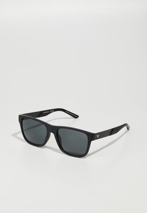 Óculos de sol retangulares pretos com lentes escuras, acabamento mate e uma marcação subtil nas hastes. A armação apresenta um design robusto.