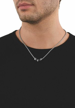 Primer plano de la parte inferior del rostro y cuello de un hombre que lleva un collar de cadena plateada con tres anillos negros y plateados sobre una camiseta negra.