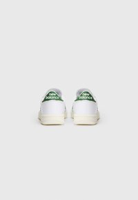 Baltas sporta apavi ar zaļiem akcentiem, ar apaļu purngalu, plakanu zoli un teksturētu virsu. "New Balance" logo redzams uz papēža.
