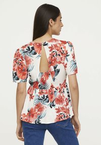 Haut floral sans dos avec des manches courtes, fond blanc, motif floral rouge et bleu, taille peplum, et ouverture en forme de clé au dos.