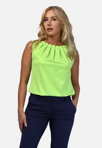 Narzucony top w jaskrawym limonkowym kolorze z plisowanym dekoltem, w połączeniu z granatowymi spodniami z małym białym wzorem w kropki.