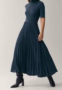 Kvinna klädd i en marinblå plisserad midiklänning med armlängd till armbågen och svarta stövletter med klack, stående mot en enkel bakgrund.