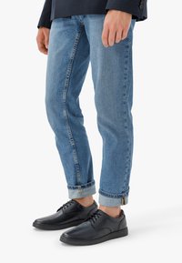 Tumši ādas apavi ar apaļiem purngaliem un šņorēm, savienoti ar sarullētām gaiši ziliem džinsiem, kuriem ir viegla noberzuma un kontrastējošā šūšana.