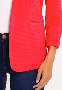 Blazer côtelé rouge vif avec une poche avant unique, manches à revers, associé à un jean bleu foncé avec un détail de bouton en cuivre.