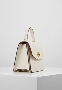 Krämfärgad läderhandväska med en strukturerad design, guldfärgad hårdvara, ett handtag ovanpå och svart kantning. Har en fällbar stängning på framsidan.