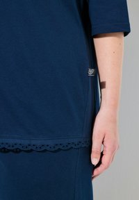 Tmavě modré bavlněné šaty s krajkovým lemem ve tvaru vlny, krátkými rukávy a detailem postranní etikety, zobrazující blízký pohled na strukturovanou látku.