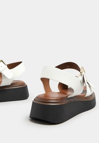 Sandali bianchi in pelle con una suola piattaforma nera spessa, dotati di un elegante fiocco laterale e cinturino regolabile alla caviglia, che mostrano una texture liscia.