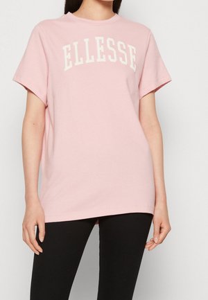 Kobieta w jasnoróżowej koszulce z krótkim rękawem z napisem „ELLESSE” i czarnych spodniach, stojąca na jednolitym jasnym tle.