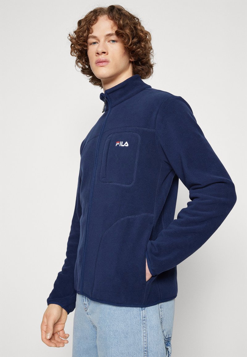 Granatowa polarowa kurtka z wysokim kołnierzem, zamek z przodu i kieszeń na piersi. Posiada logo na kieszeni oraz gładką teksturę.