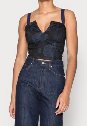 Femme portant un crop top floral foncé à larges bretelles et un jean bleu foncé taille haute, debout devant un fond clair uni.