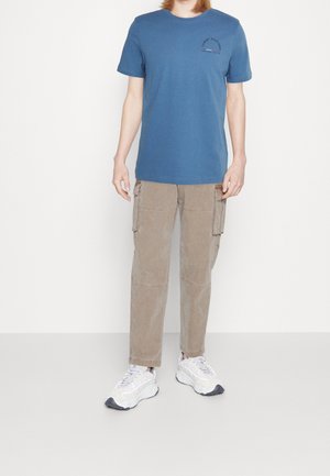 Niebieska bawełniana koszulka z małym nadrukowanym logo na klatce piersiowej, zestawiona z beżowymi spodniami typu cargo z wieloma kieszeniami. Na nogach białe trampki.
