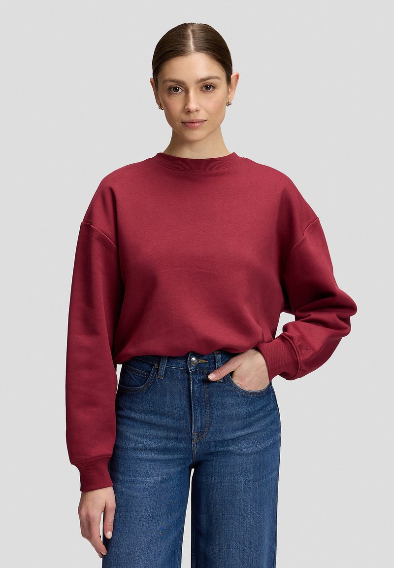 Jeune femme aux cheveux attachés, portant un pull rouge à manches longues et un jean bleu taille haute, debout devant un fond uni.