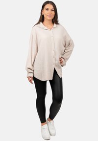 Beige button-up shirt met wafelstructuur, met kraag, lange mouwen en een gebogen zoom, gecombineerd met zwarte leggings en witte sneakers.