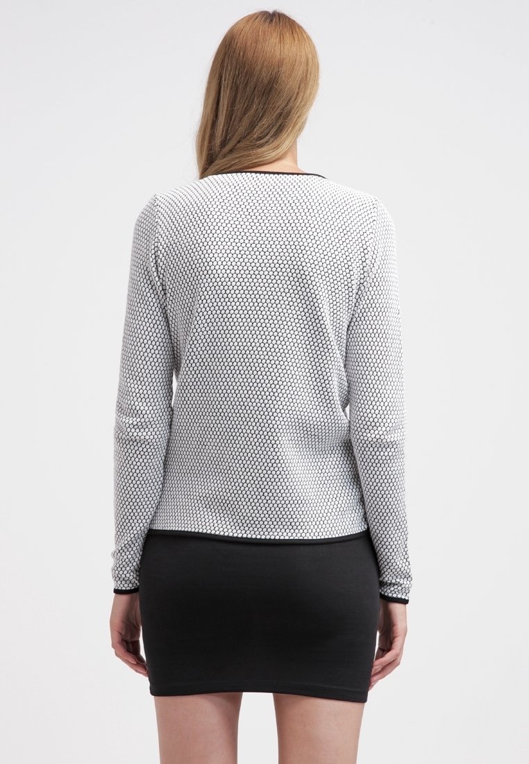 Femme aux longs cheveux blonds portant un haut blanc à manches longues avec un motif nid d'abeille et une jupe noire ajustée, debout de dos.
