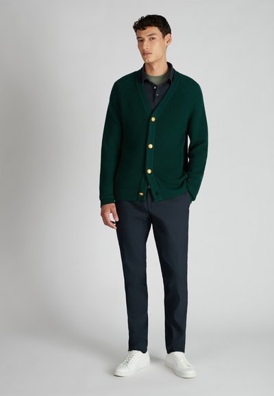 Homme debout portant un cardigan boutoné vert foncé, des chemises noires et vertes superposées, un pantalon bleu marine et des baskets blanches sur fond gris uni.