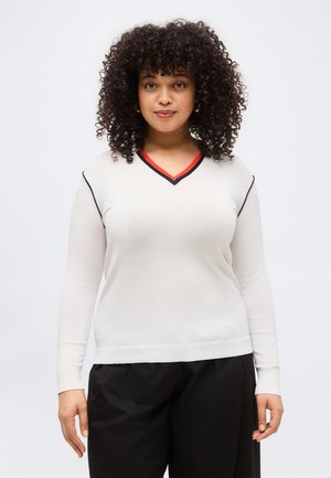 Frau mit lockigem Haar, die einen weißen V-Ausschnitt-Pullover mit schwarzem und rotem Besatz sowie eine schwarze Hose trägt, vor einem schlichten weißen Hintergrund steht.