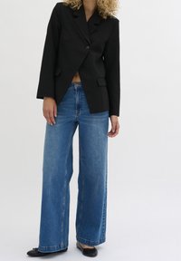 Czarna marynarka na jedno zapięcie z ostrymi klapami, zestawiona z jasnoniebieskimi szerokimi spodniami jeansowymi. Oba elementy wykonane z tkaniny o gładkiej fakturze.