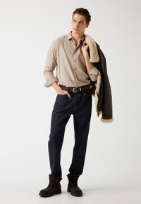 Beige button-up shirt, donkerblauwe jeans, bruine riem en bruine veterschoenen. Een zwarte jas met een beige bontvoering hangt over zijn schouder.