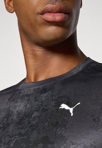 Svart atletisk skjorte med teksturmønster, rund hals, og hvit Puma-logo på øvre venstre bryst. Glatt stofffinish.