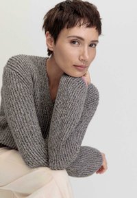 Grauer gestrickter Pullover mit V-Ausschnitt, strukturiertem Muster und langen Ärmeln. Das Model posiert mit einer Hand unter dem Kinn vor einem neutralen Hintergrund.
