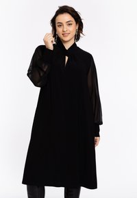Robe noire avec un col montant, un détail en trou de serrure et des manches longues transparentes. Le tissu semble lisse avec une silhouette fluide, atteignant le mollet.